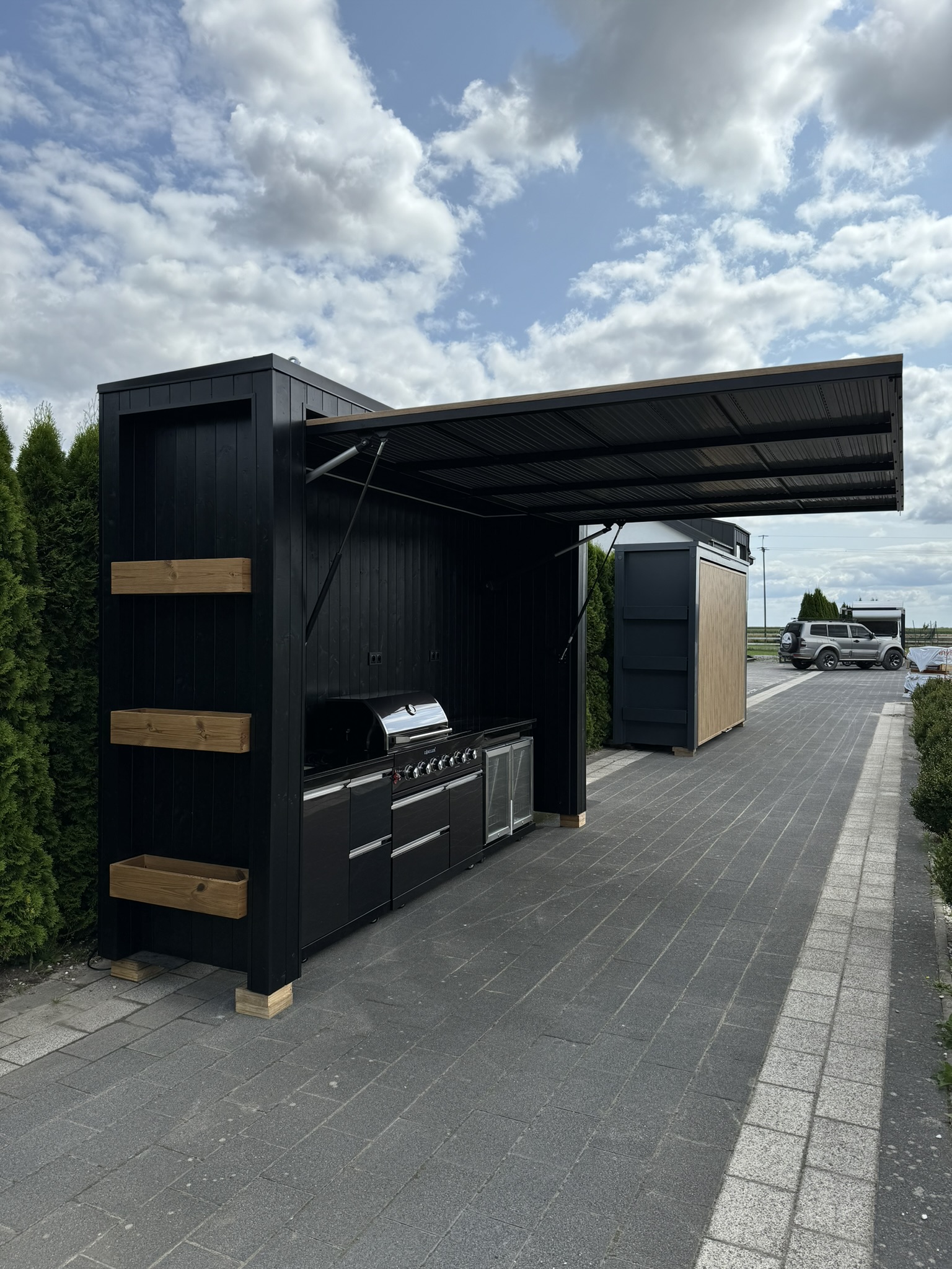 Black outdoor kitchen with a lift-up canopy, gas barbecue and fridge on block paving.