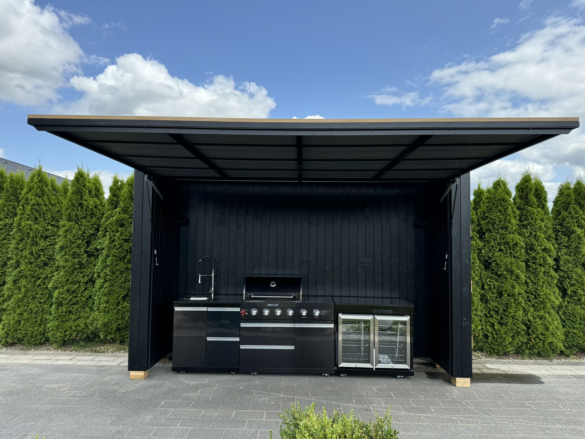 Black outdoor kitchen with an open canopy, equipped with a barbecue, sink and glass-fronted fridge, set against a backdrop of thuja trees.
