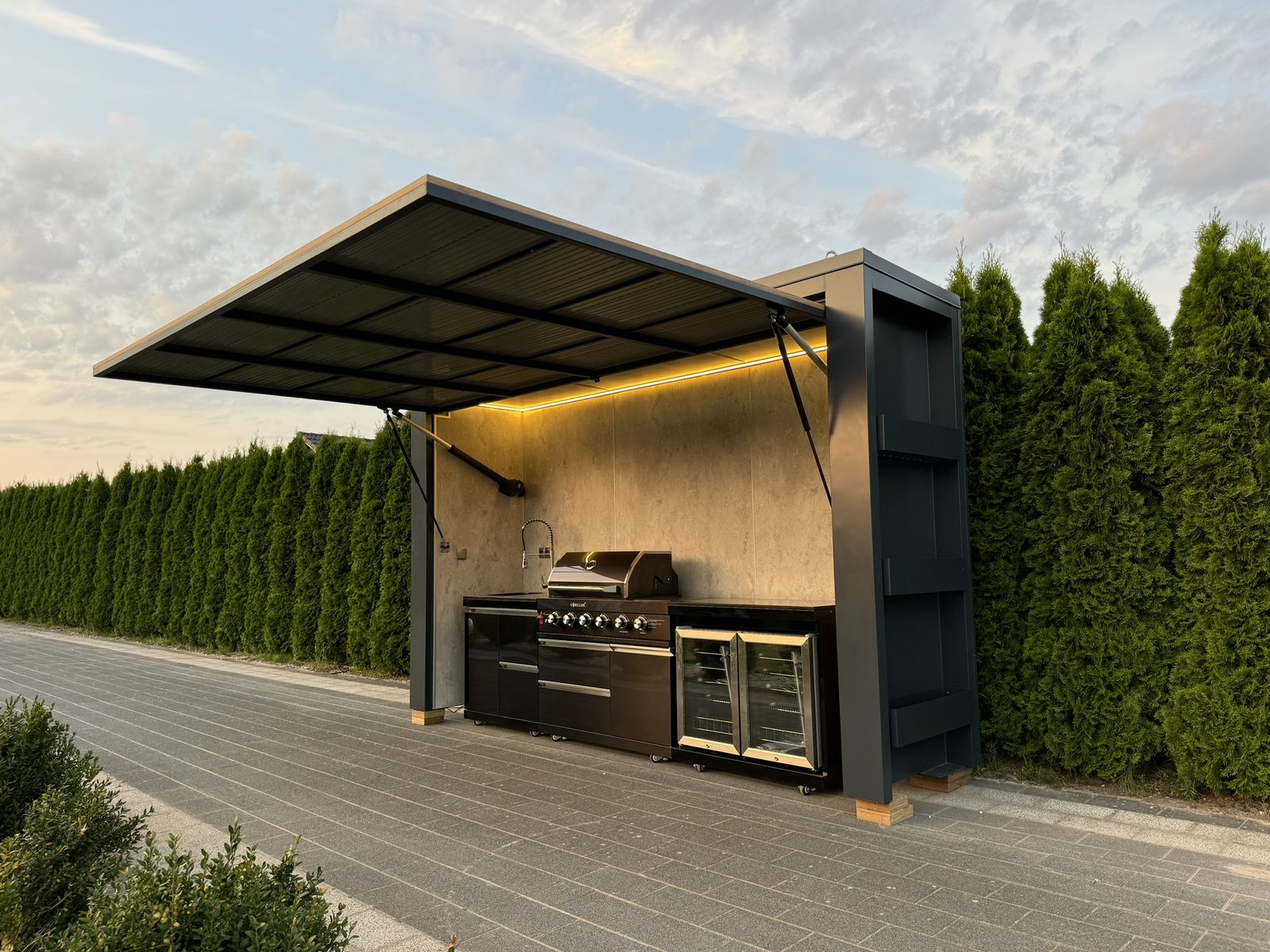 Black open outdoor kitchen with an LED-lit canopy, barbecue and fridge, set against a hedge.