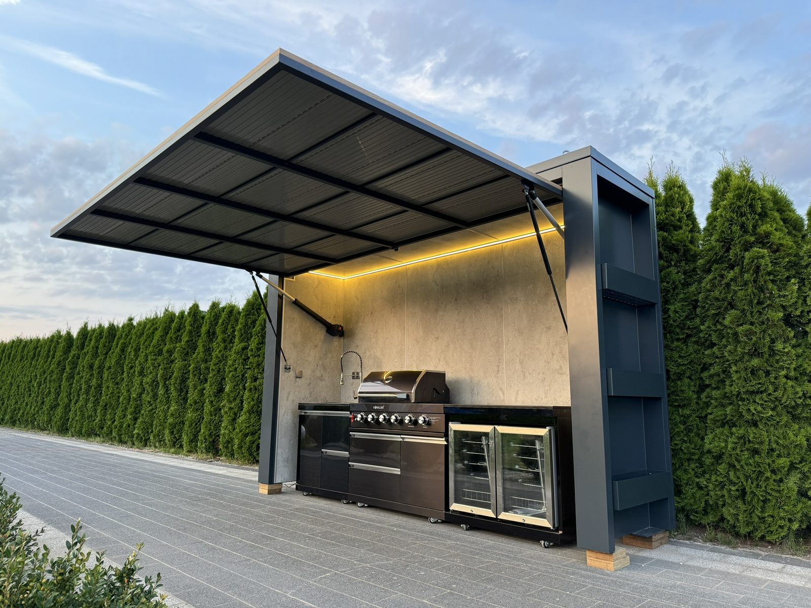 A modern outdoor kitchen with an open LED-lit canopy, barbecue, sink and fridges beside a hedge.