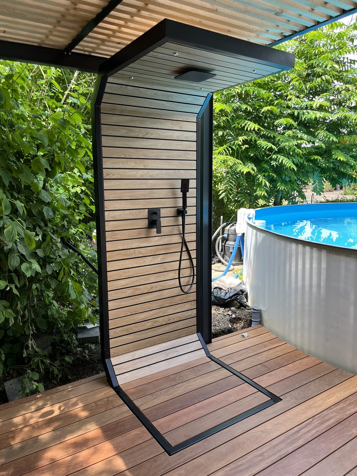 Modern outdoor shower with wooden slats and black fittings on a terrace beside a swimming pool.