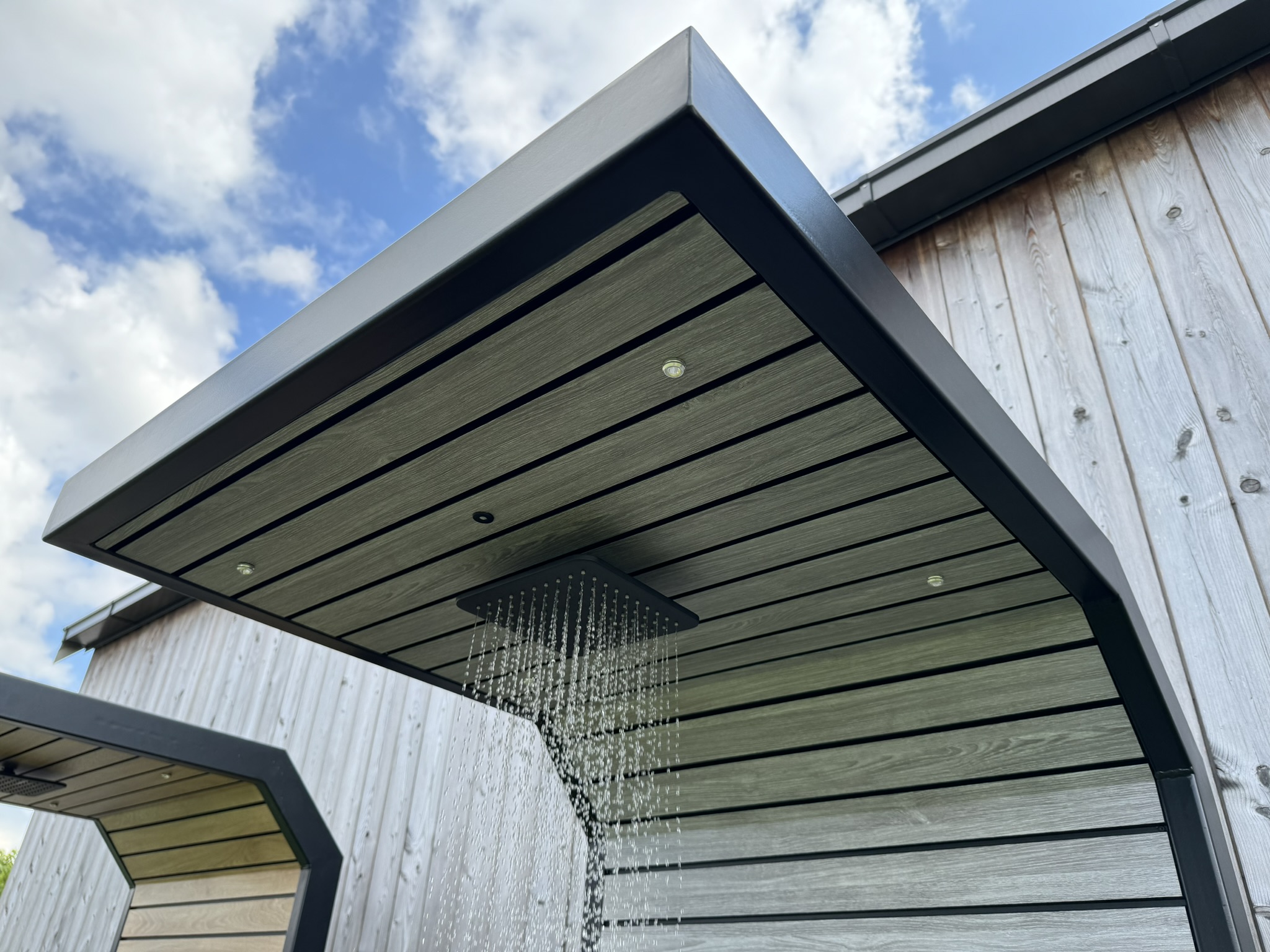 Freestanding outdoor shower with a rainfall showerhead running water and spot lighting in the canopy.
