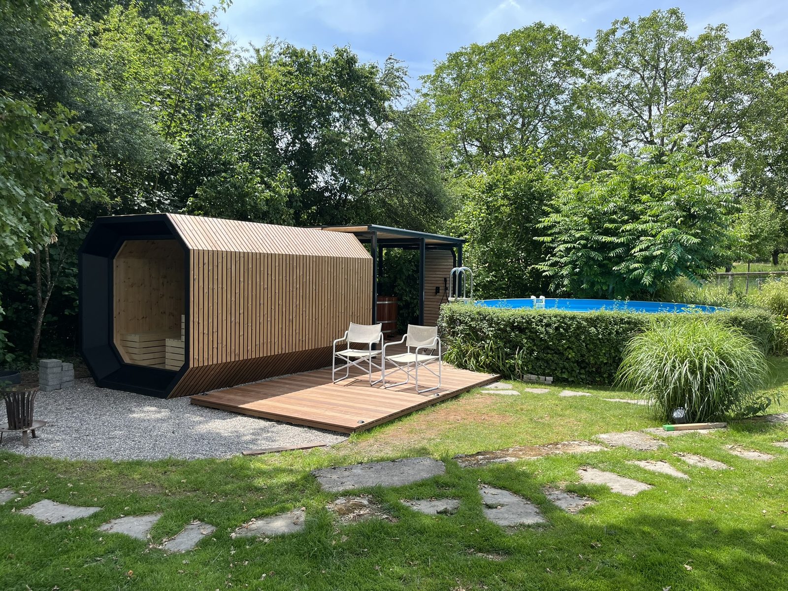 A modern garden sauna clad in wooden slats, set on a wooden terrace in a garden beside a swimming pool.