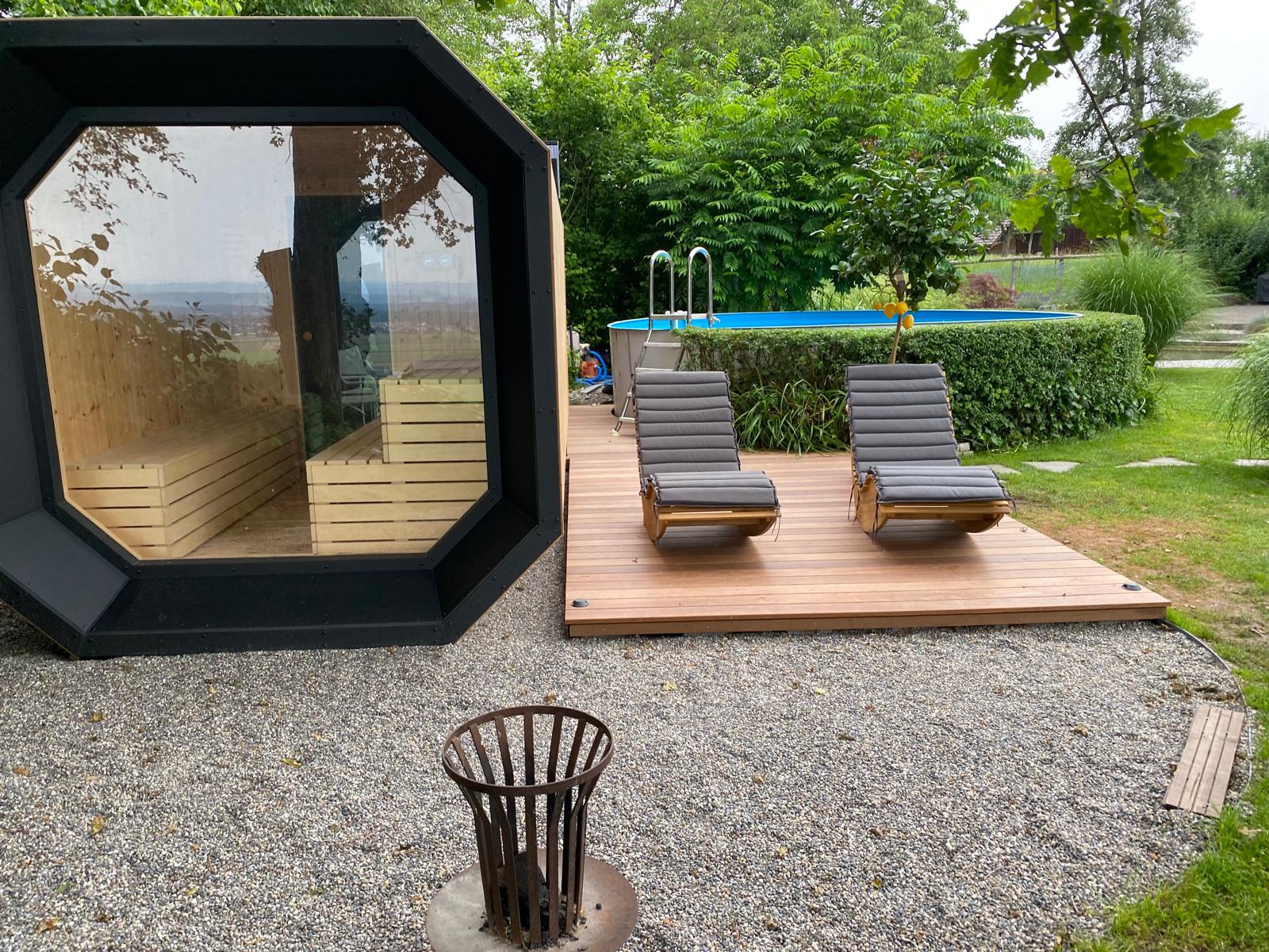 A modern garden sauna with a glazed front beside a wooden terrace with sun loungers and a swimming pool.