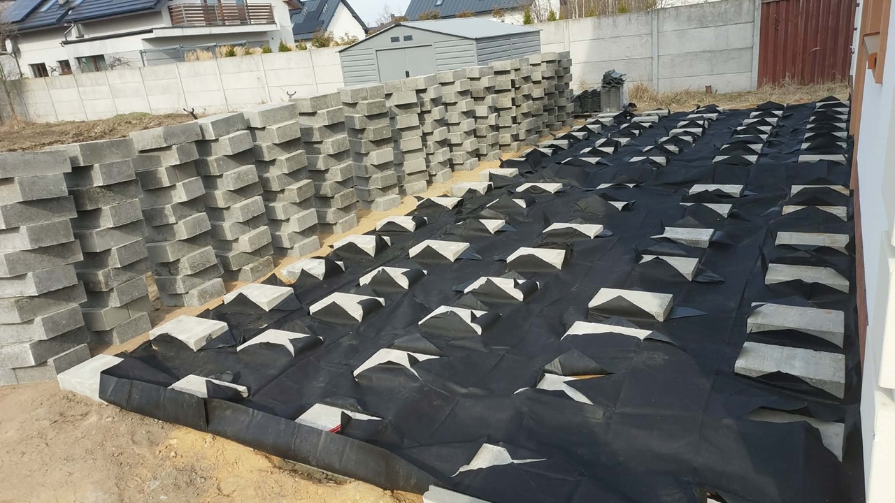 Preparing the terrace base: concrete blocks laid on black geotextile beside a building.