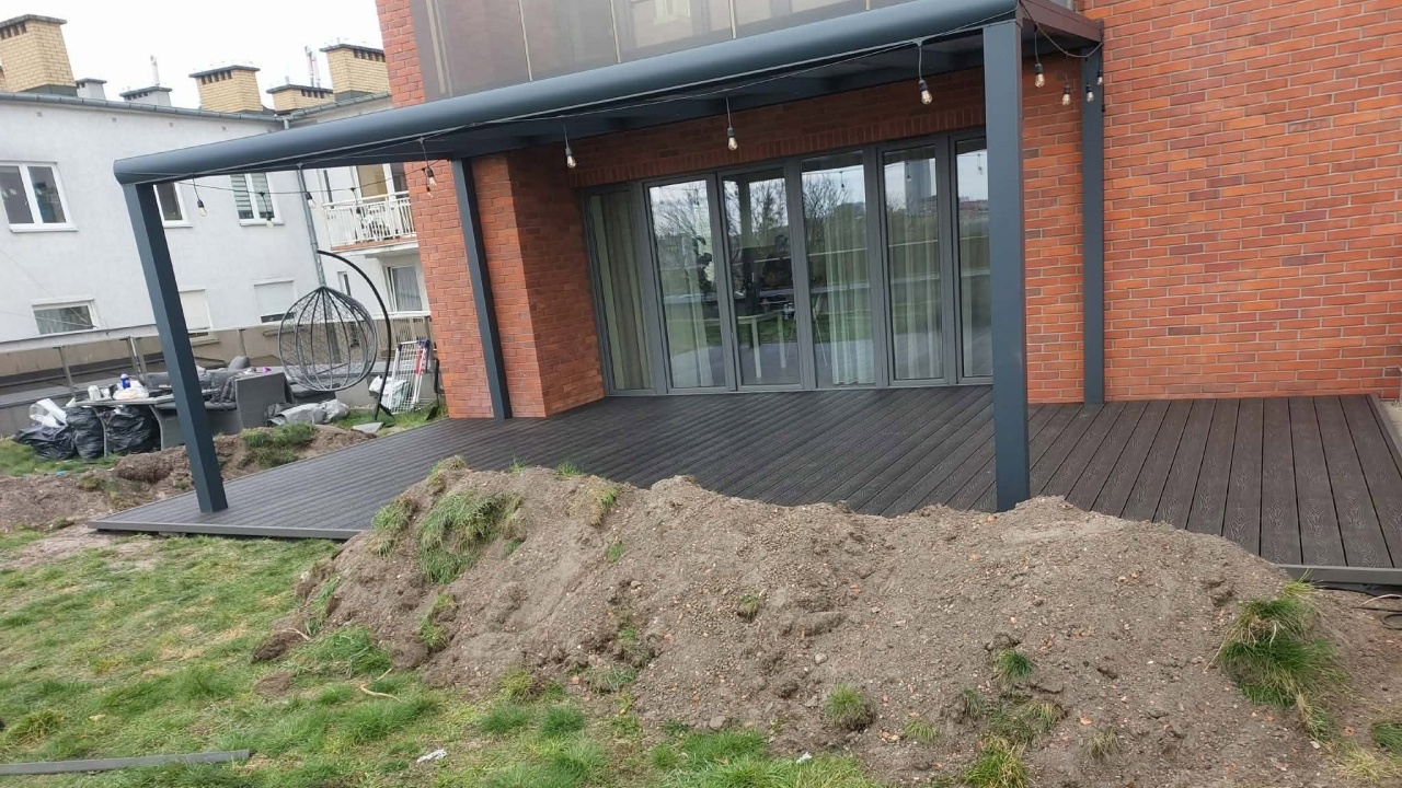 A terrace made from graphite-grey composite decking boards under a dark canopy beside a red-brick building.