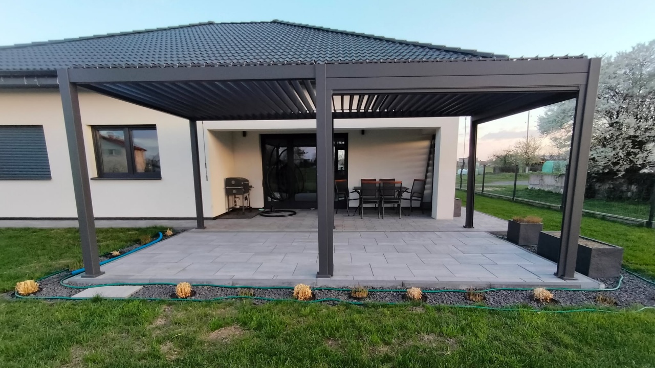 Dark grey louvred pergola sheltering a grey patio beside a detached house, with garden furniture.