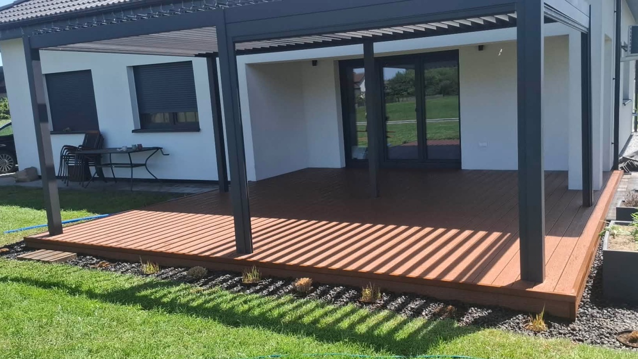 A terrace made from copper-coloured composite decking boards under a graphite-grey pergola beside the house façade.