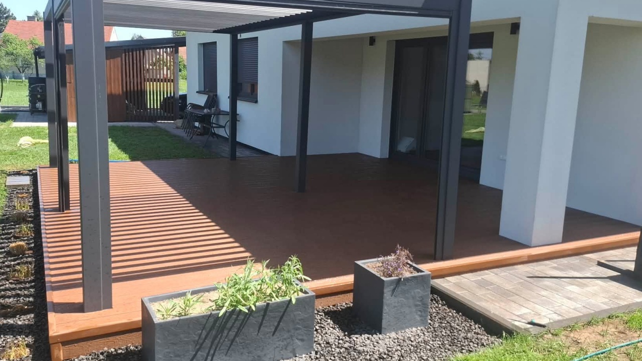 Modern terrace made of copper-coloured composite decking boards beside a house, with a black pergola and planters on gravel.