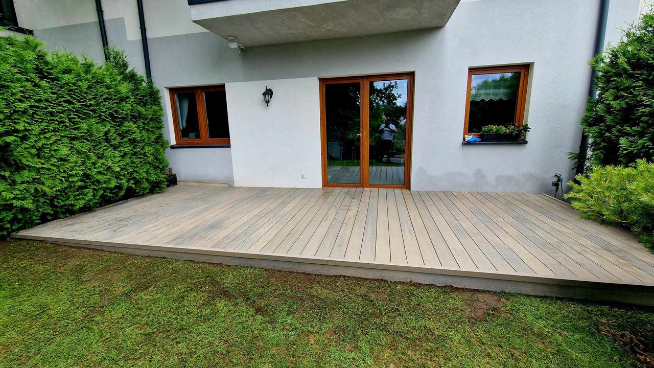 A light grey composite decking board terrace installed beside a house, surrounded by lawn and a green hedge.