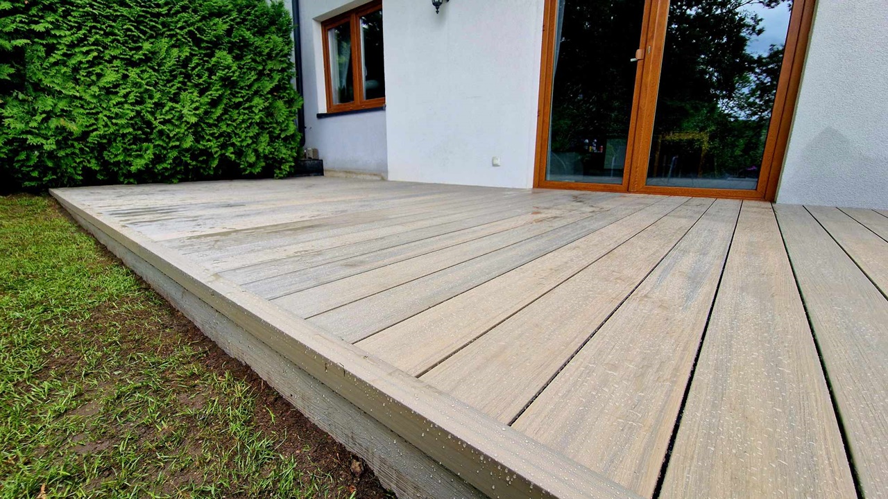 A terrace made from light brown PREMIUM composite decking boards beside a building façade, next to a lawn and hedge.