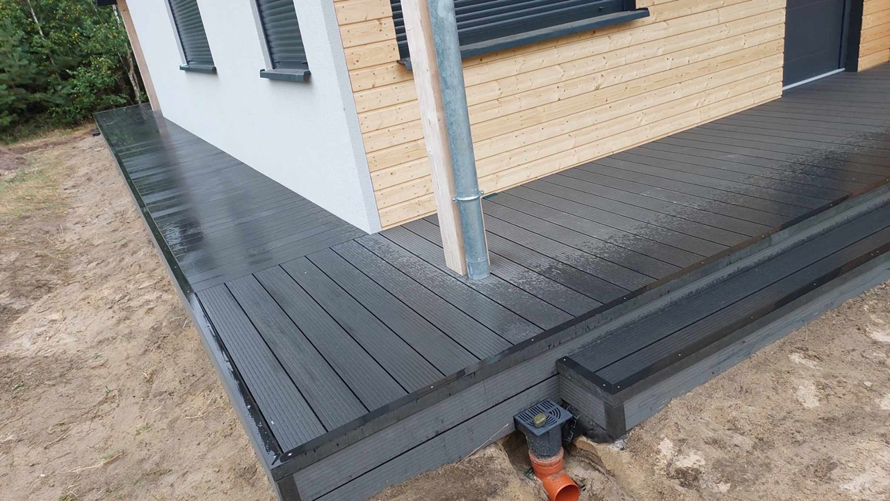 A corner terrace made of dark grey composite decking boards, with a step beside a building with timber cladding.