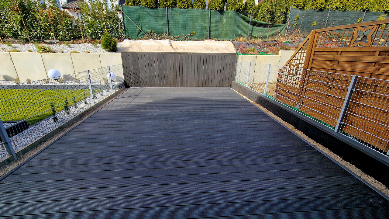 A terrace made from dark grey composite decking boards in a garden beside a house, with panel fencing.