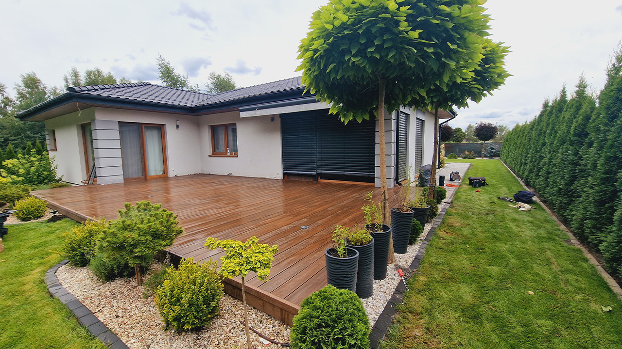 Large brown composite decking terrace beside a modern single-storey house, with a well-kept garden and plants.