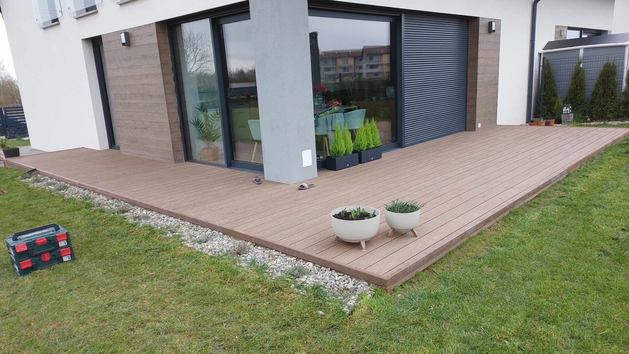 Modern terrace made of brown composite decking boards built beside a detached house with large windows.